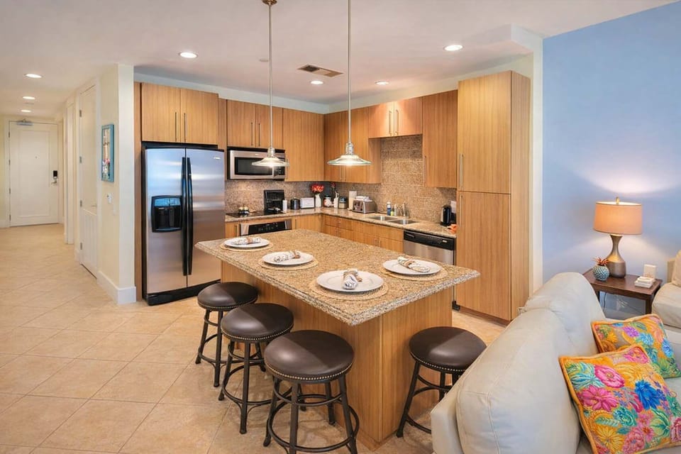 Modern condo kitchen with granite island, stainless steel appliances, and cozy living area—perfect for families or couples. Steps from the beach, fully stocked, and ideal for relaxing getaways or remote work stays.