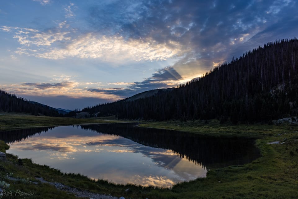 A serene lake reflects the sky at sunset, surrounded by green grass and silhouetted trees against a backdrop of darkening mountains and scattered clouds.