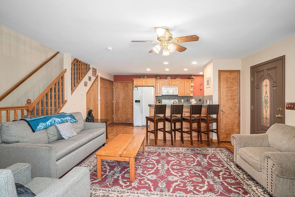 Living room with gray sofas, wooden coffee table, patterned rug, ceiling fan, bar stools, and view of a kitchen with wooden cabinets and a refrigerator. Stairs lead to an upper floor.