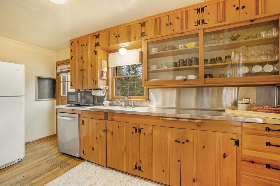 A kitchen with wooden cabinetry, a white refrigerator, a dishwasher, microwave, and a window above the sink. Upper cabinets have glass doors displaying dishes and glasses.