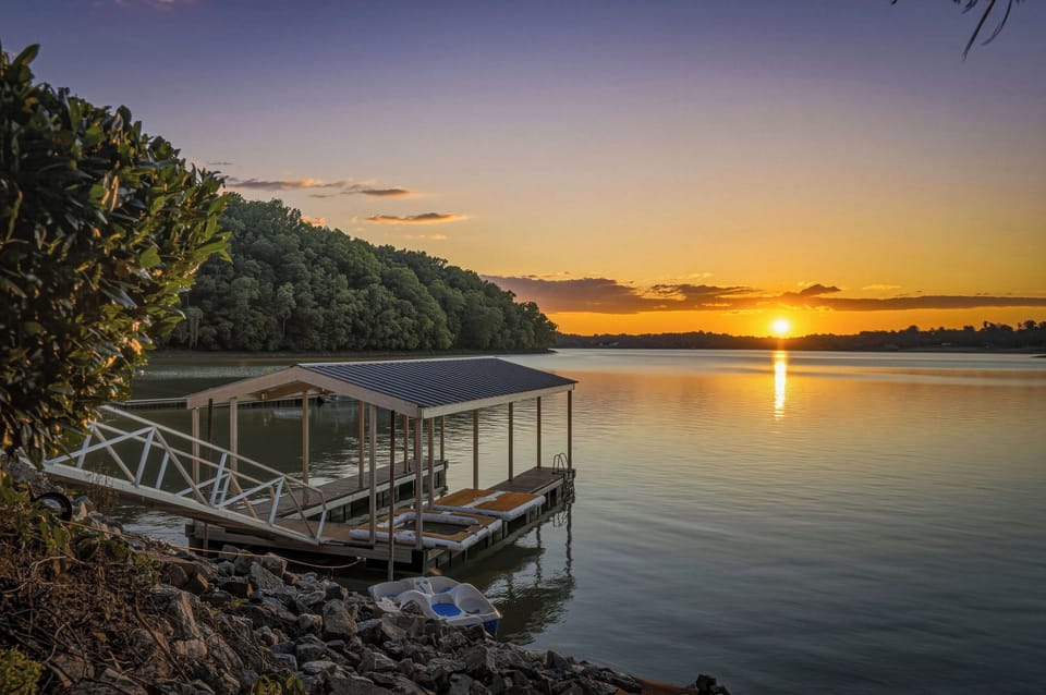 A tranquil lakeside setting features a covered boat dock against a stunning sunset, surrounded by lush forests and calm waters reflecting the evening sky.