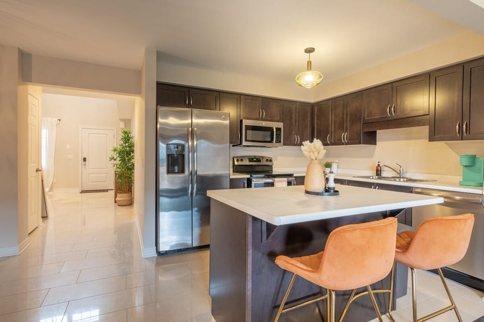 Modern kitchen with stainless steel appliances, dark wood cabinetry, and a spacious island. Bright lighting complements the open, airy design, while the stylish bar stools add a touch of warmth. Perfect for cooking and gathering with loved ones!