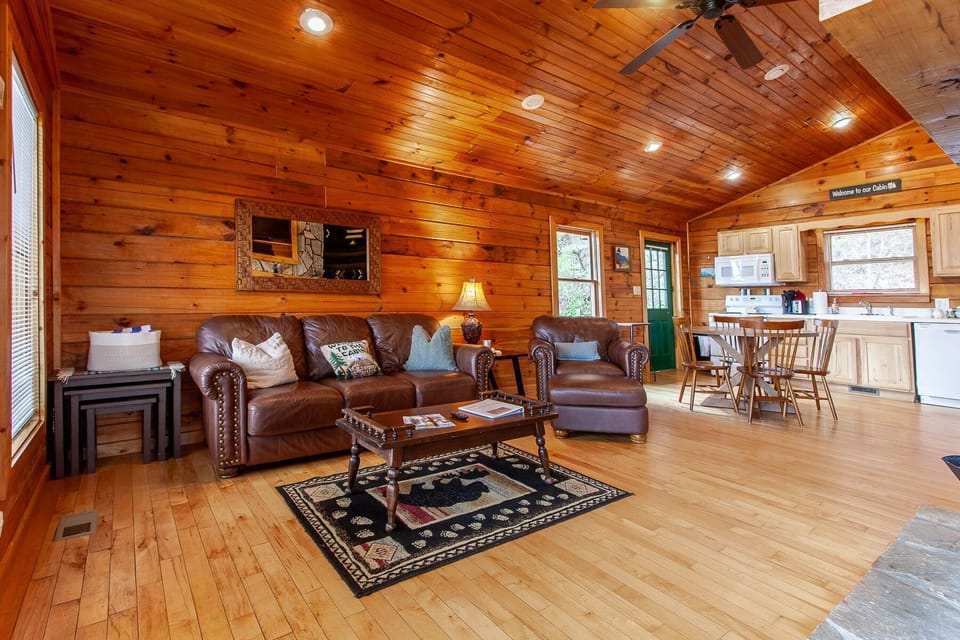 Open concept great room with leather seating, wood interior walls, vaulted ceiling, dining area, and kitchen visible in the background. This inviting cabin layout in Bryson City offers a spacious gathering space with mountain cabin charm.