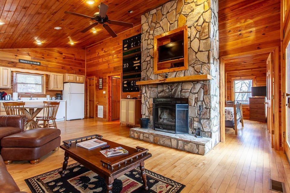 Great room featuring a floor-to-ceiling stone fireplace, leather furnishings, wood-paneled walls, and open access to the kitchen. The spacious interior highlights the cozy mountain cabin atmosphere near the Smoky Mountains.