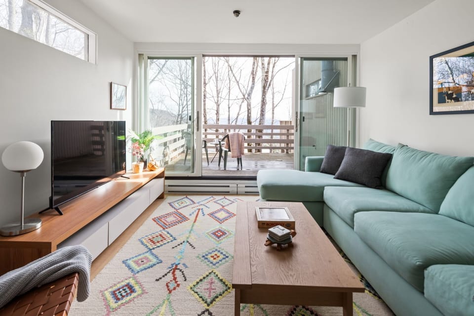 - Spacious living room with sectional sofa and balcony access   - Streaming ready TV and vibrant rug add comfort to Airbnb stay   - Vacation rental with indoor lounge and outdoor forest backdrop