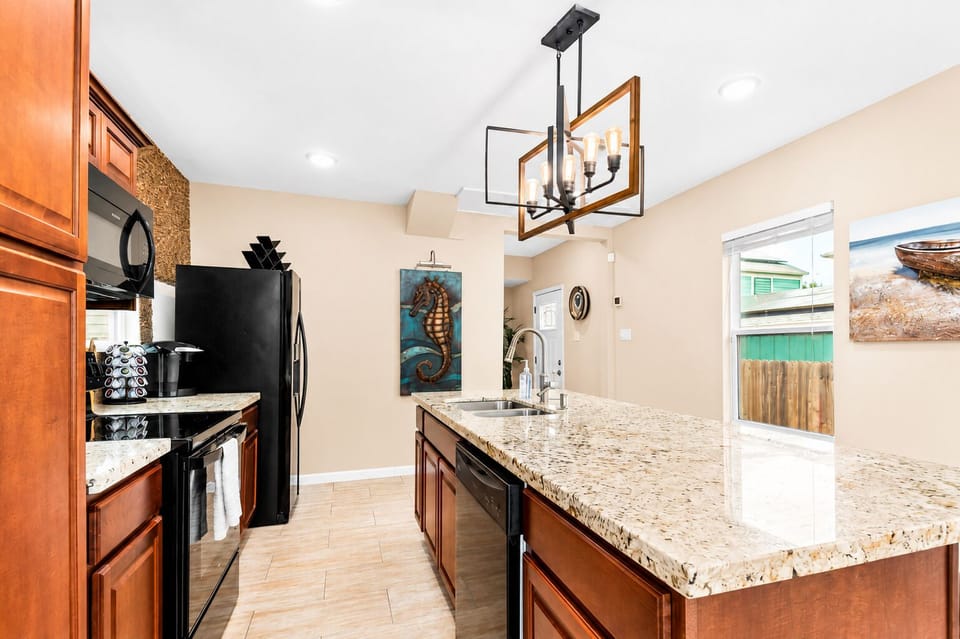 Galley-style kitchen with granite island, double sink, wood cabinetry, black refrigerator, microwave and stove, plus access to the backyard.