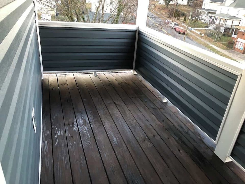 The residential balcony offers a private outdoor space with dark-stained wooden flooring and high metal siding, providing a secluded area for relaxation with a view of the neighborhood.