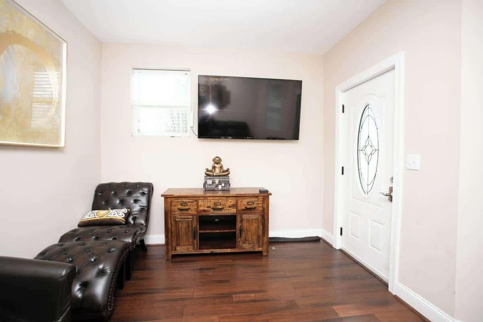 The living area features a tufted leather chaise lounge and a rustic console table beneath a wall-mounted flat-screen TV. A large abstract painting adds a touch of sophistication, while the entry door with decorative glass offers a warm welcome.