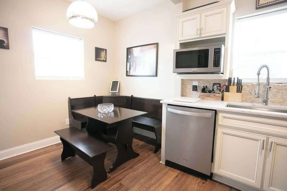 The kitchen dining space features a sleek, dark wood corner nook with a matching table under a chic pendant light. Cream cabinetry, a microwave, and modern appliances complement the mosaic backsplash and wooden flooring, creating a harmonious design.