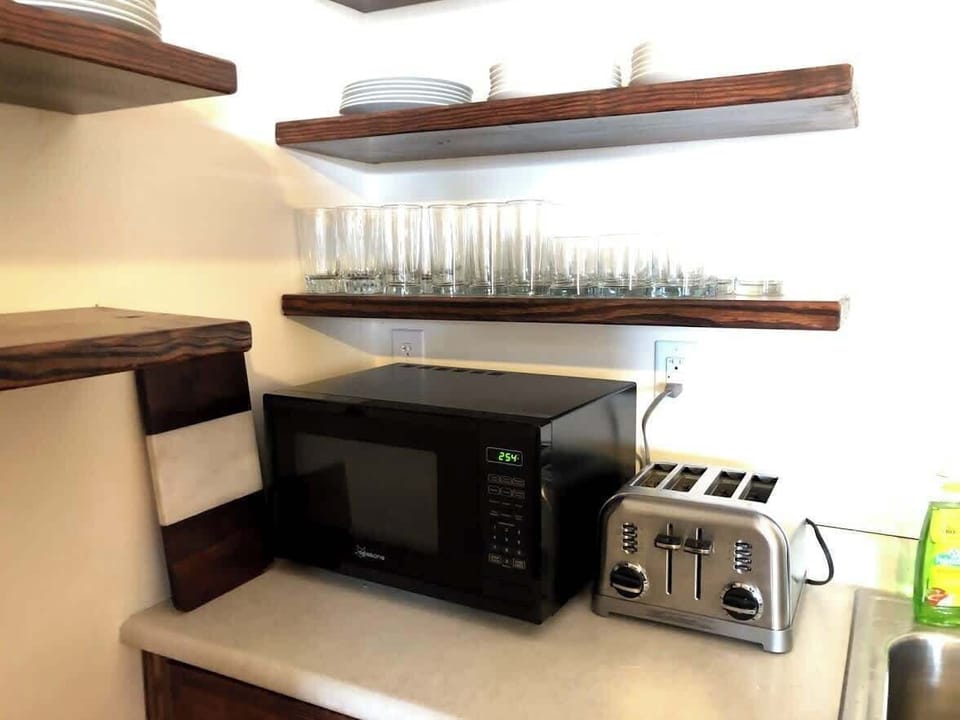 The kitchen corner is practically laid out with a microwave and four-slice toaster on the countertop. Wooden shelves above hold a neat row of glasses, contributing to the space's tidy and efficient design, suitable for quick meal preparations.