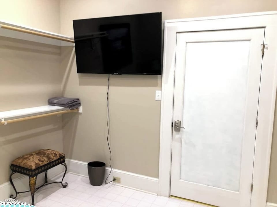 A modern setup with a flat-screen TV elegantly mounted on the wall adjacent to a white closet, which doubles as a bench for added functionality. A sleek black trash can discreetly completes the tidy arrangement.