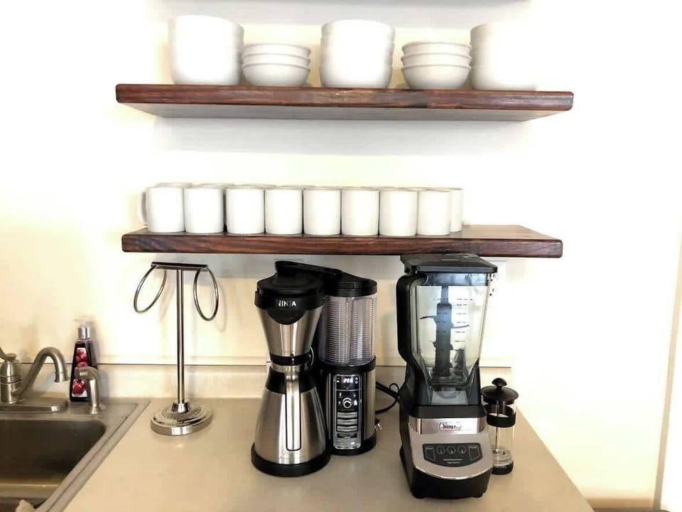 The kitchen counter is neatly organized with a coffee maker and a blender, ready for use. Above, open wooden shelves hold a clean array of white bowls and mugs, emphasizing the kitchen's practical and minimalist aesthetic.