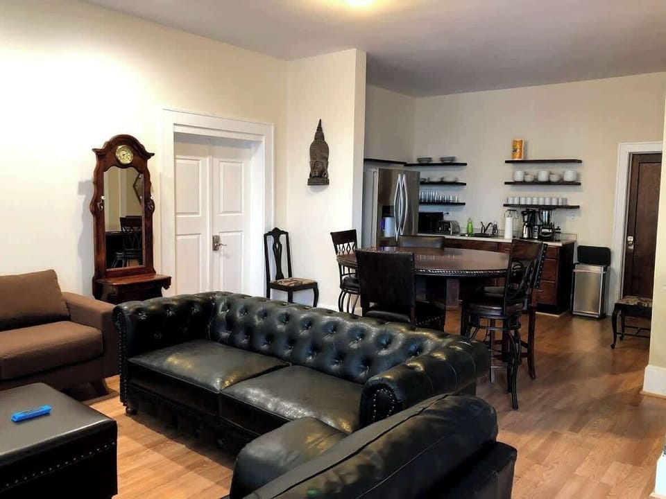 The living room is comfortably furnished with a brown sectional sofa and a tufted leather ottoman. A large window draped with sheer curtains allows natural light to complement the warm tones of the room.