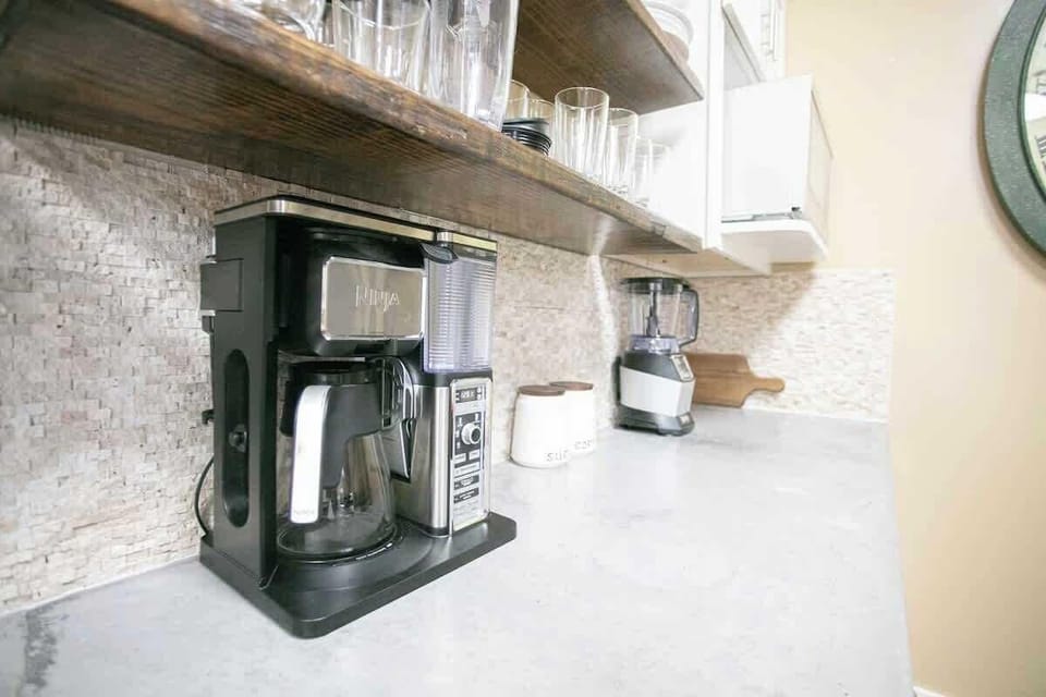 The kitchen counter is neatly organized with a modern coffee machine and a blender, ready for use. Above, open shelving displays a variety of glasses, showcasing a functional yet stylish setup.