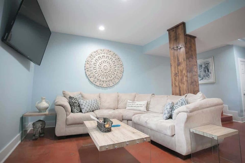 The living area is cozy with a large, comfortable sectional sofa adorned with decorative pillows. An intricate wall mandala, a rustic column, and a sleek wall-mounted TV contribute to the room's modern yet homely charm.