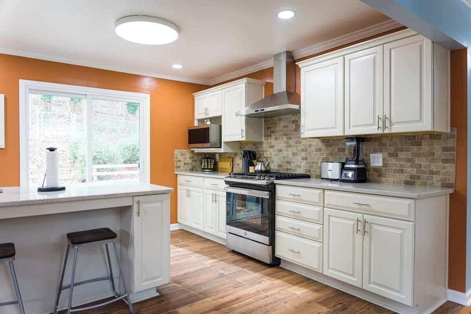 The kitchen is warmly lit and welcoming, featuring cream cabinets, a tiled backsplash, and modern appliances. A large window invites natural light, complementing the earthy tones and hardwood flooring for a cohesive, inviting space.