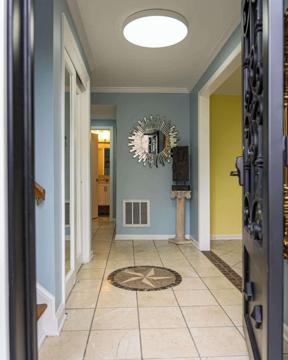 The photograph depicts a spacious hallway with light blue walls and a large, decorative sunburst mirror as the focal point. The floor features a neutral tile with a detailed compass rose design inlay, leading towards a warmly lit kitchen in the background