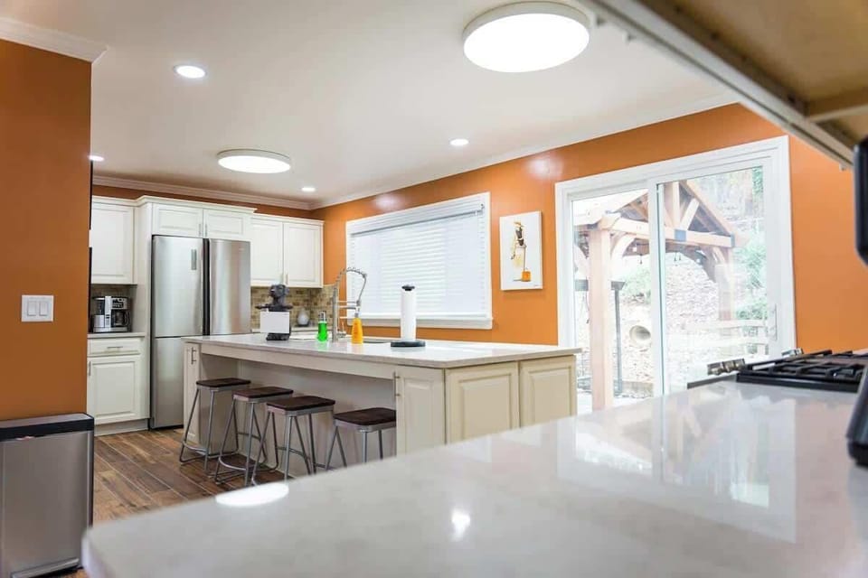 The image captures a spacious and modern kitchen with rich terracotta walls and elegant white cabinetry. The room is well-lit by recessed ceiling lights and natural light from a window overlooking an outdoor pergola.