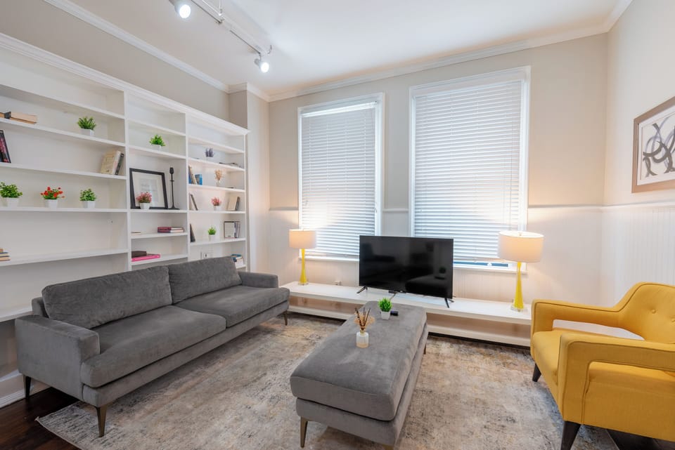 Relax in this light-filled living room featuring a plush sofa, cheerful accent chair, and built-in bookshelves — the perfect spot to unwind with your favorite show or a good book.