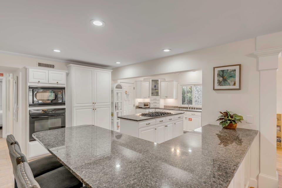 A modern kitchen with white cabinets, a built-in oven, a gas stove, and a large granite island. A framed picture hangs on the wall next to a small potted plant.