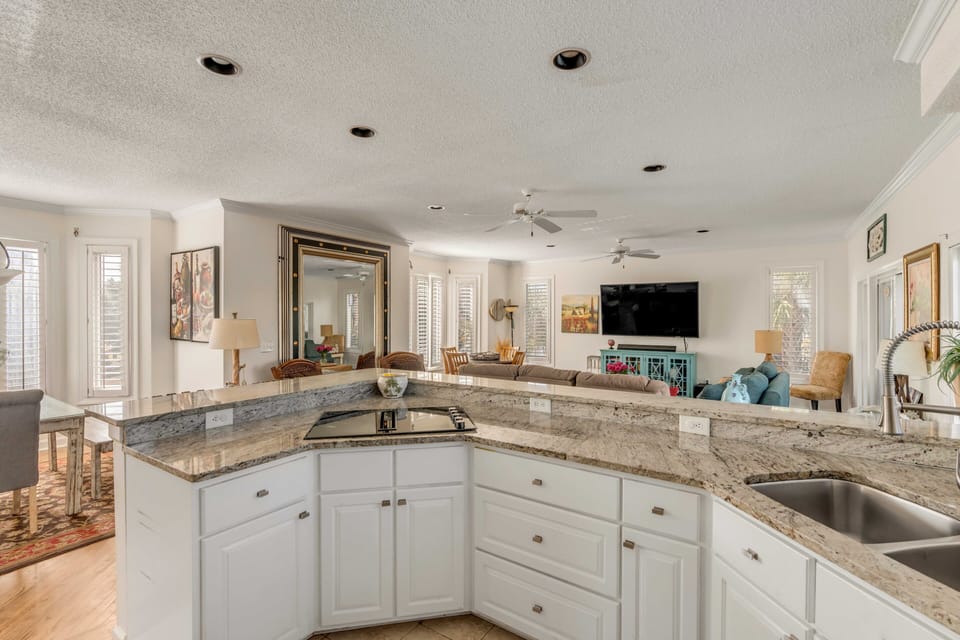 Spacious open-concept living area with a kitchen island, granite countertops, white cabinets, a dining table, and a seating area facing a large TV and artwork on the walls.