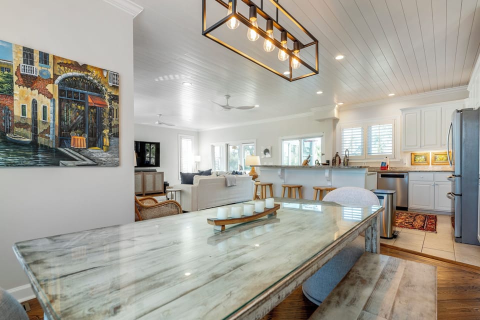 A dining room with a rustic table, benches, and gray chairs. Large windows with shutters provide natural light. A pendant light hangs above the table, and artwork decorates the walls.