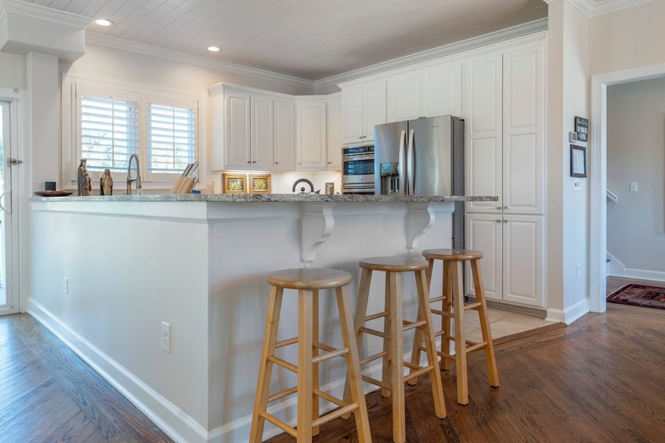 Bright kitchen with white cabinets, stainless steel appliances, and a granite countertop. Four stools line the bar, while a doorway and windows allow natural light to fill the space.