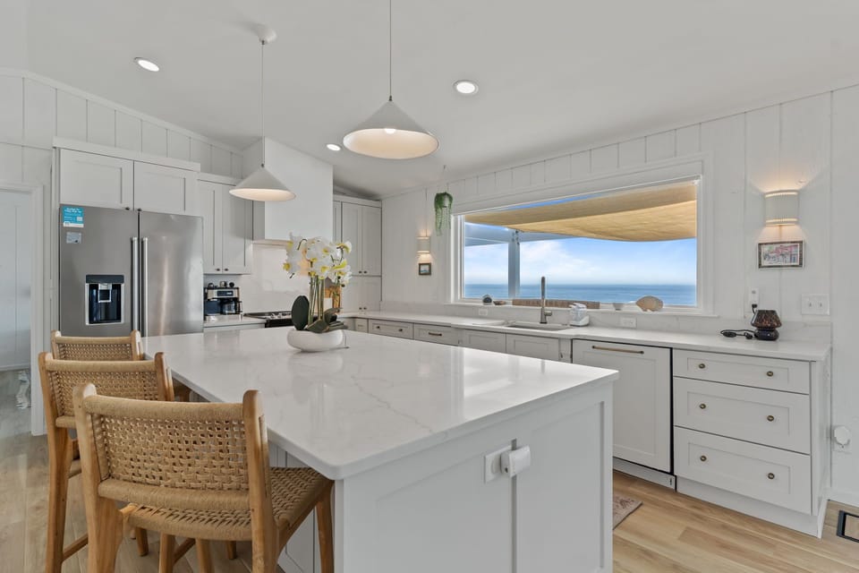 Bright white kitchen with a large island, quartz counters, stainless appliances, farmhouse sink, and a big window overlooking the porch and ocean.