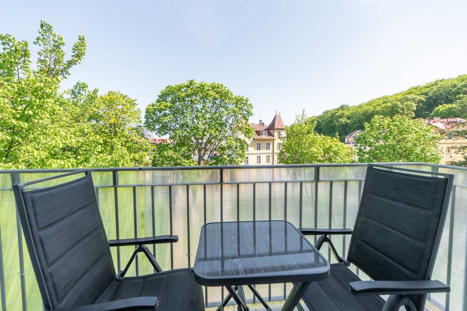 A balcony equipped with a small table and two chairs, surrounded by greenery and trees in the background, ideal for enjoying a peaceful outdoor moment.