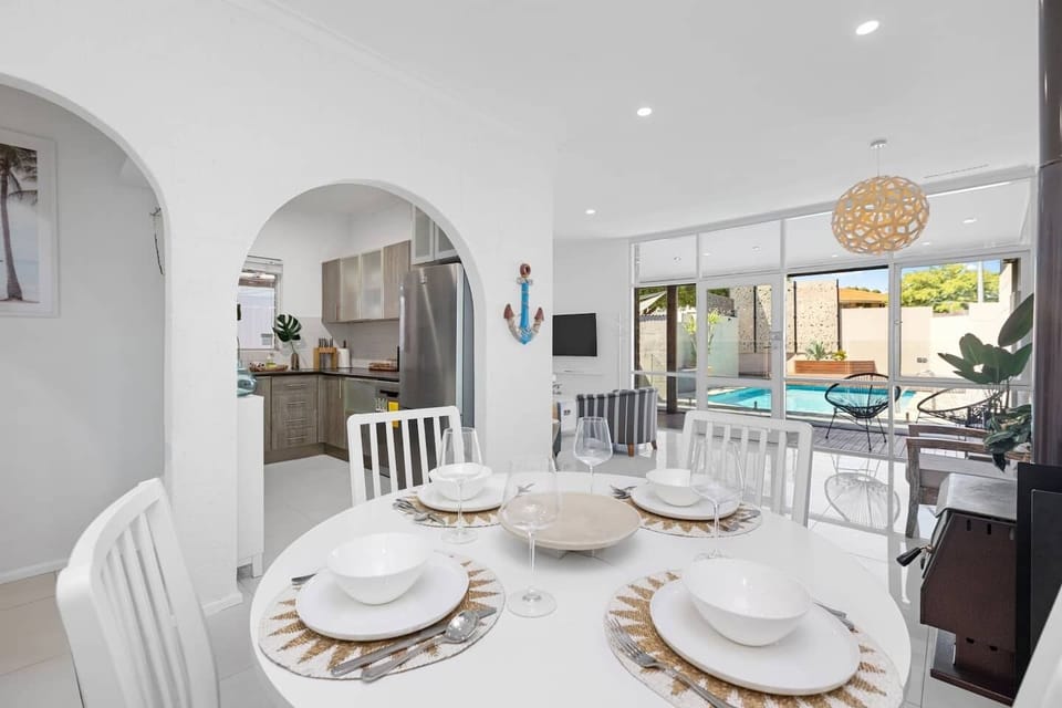 Elegant dining area with a round table, seamlessly connecting to a modern kitchen through an archway, and offering a delightful view of the outdoor pool.