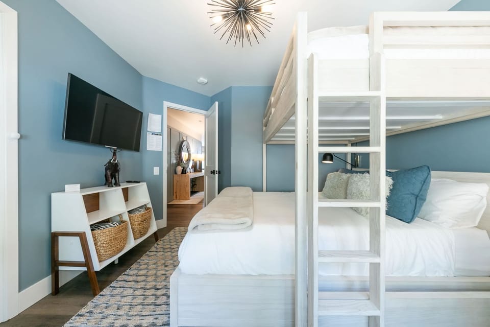 The guest bedroom is filled with natural light and includes a quiet workspace with a desk and chair tucked next to the windows, a spacious closet, and a 43” mounted smart TV.