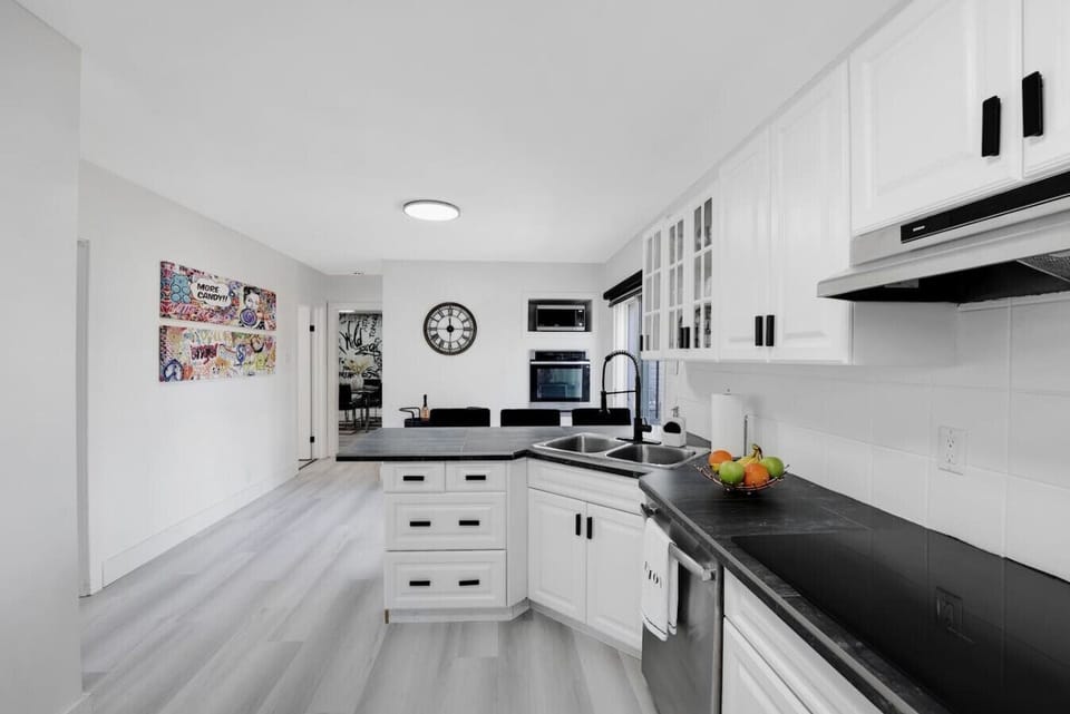 Bright, modern kitchen with sleek white cabinetry, dark countertops, and a stylish range hood. Features a central island, ample storage, and a clean, inviting atmosphere. Perfect for culinary adventures. Book your stay and enjoy this fantastic space!