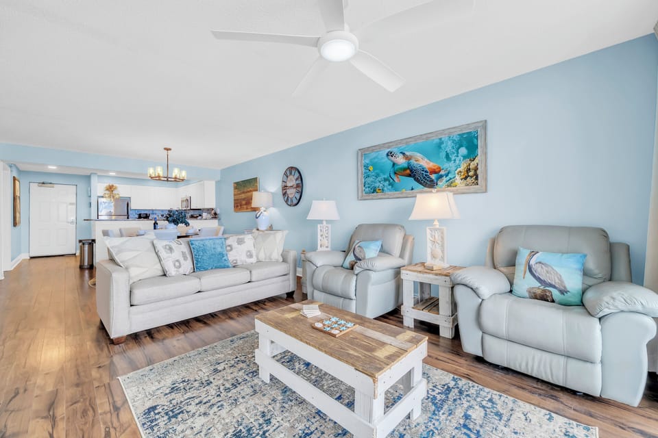 Relax in this coastal-themed living space with comfortable seating, ocean artwork, and an open layout that flows seamlessly into the kitchen area.