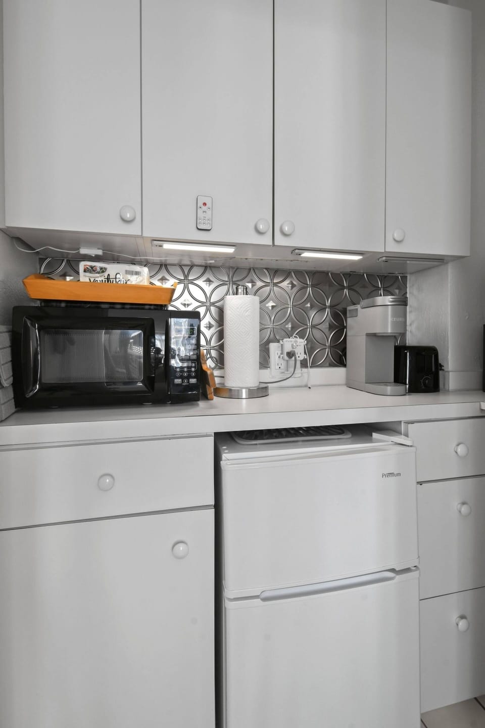 Kitchenette with microwave, toaster, mini fridge, Keurig coffee maker, glasses, plates, and cutlery. No Sink