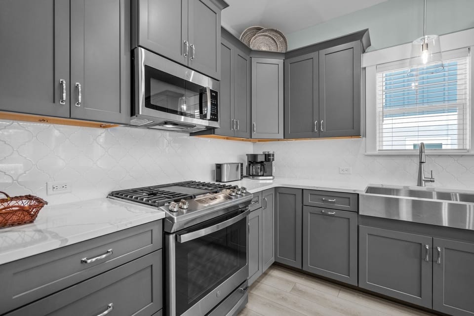 Bright kitchen with gray cabinets, stainless appliances, large island, white stools, farmhouse sink, dishwasher, microwave, coffee maker, toaster, and coastal Oak Island, NC style.