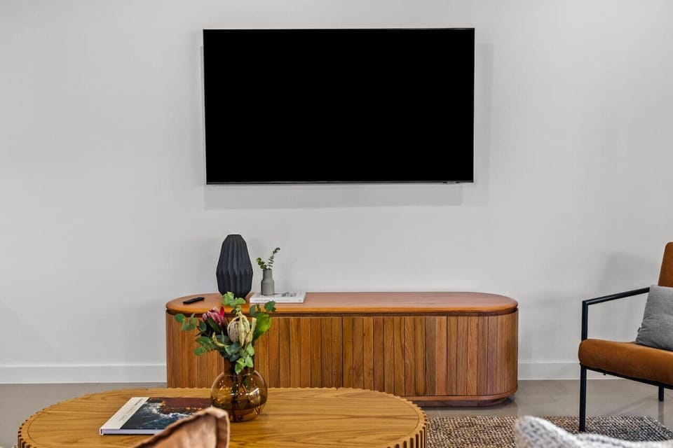 A modern living room setup with a wooden entertainment unit under a large blank TV screen, a vase with flowers on the unit, a round wooden coffee table with a magazine, and an orange chair with a gray pillow.