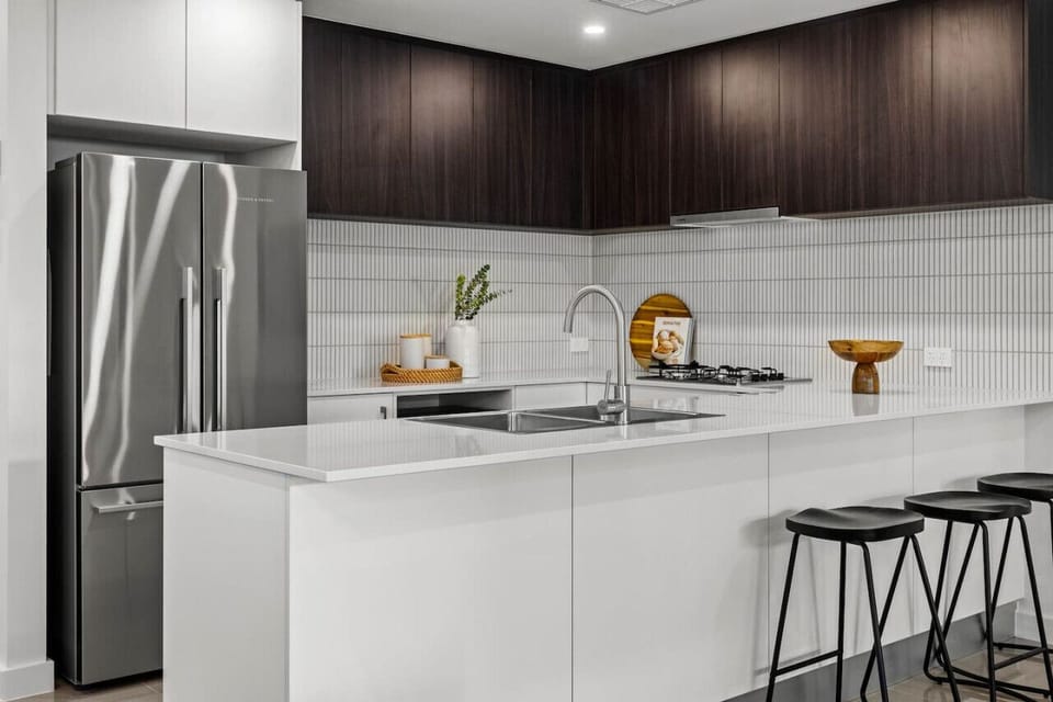 A modern kitchen with a sleek design, featuring a stainless steel refrigerator, dark wooden cabinets, white countertops, and a tiled backsplash. There are also some decorative elements like a plant and a wooden bowl, along with black stools at the counter