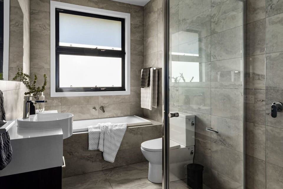 A modern bathroom with a sleek design. It features a large frosted window, a white bathtub with a towel draped over it, a white toilet, a stylish sink with a plant decoration on the counter, and gray marble tiles covering the walls, creating a clean and c