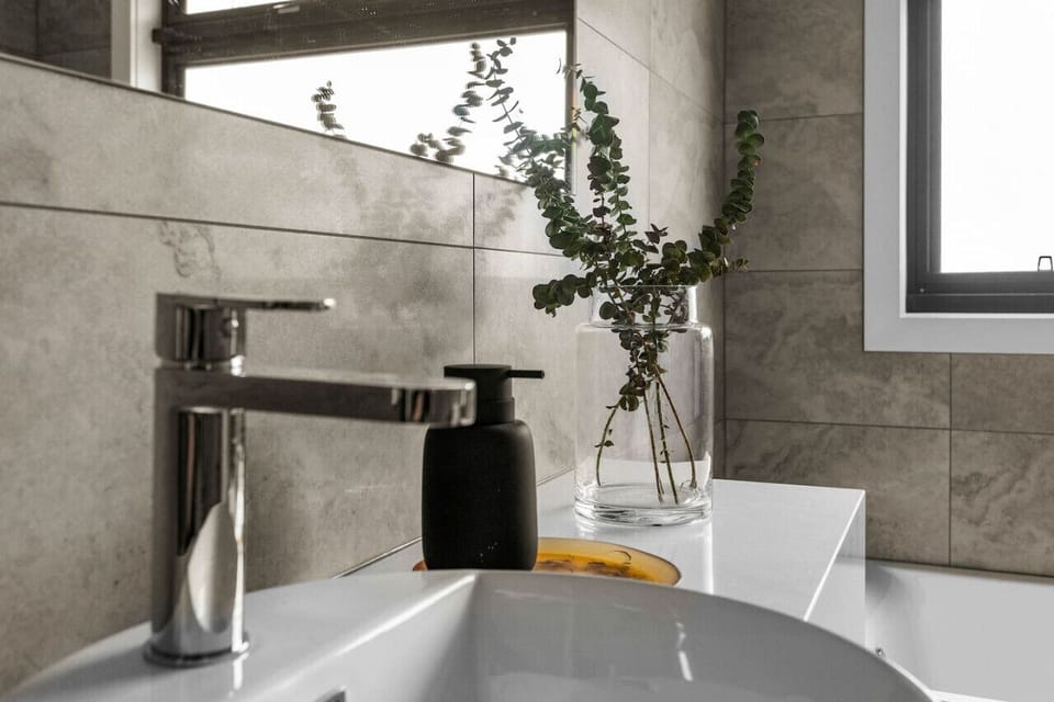 A modern bathroom sink area with a sleek chrome faucet. There is a black soap dispenser next to the faucet, and a clear glass vase with green eucalyptus branches on the countertop, adding a touch of nature to the space. The background features grey tiled 