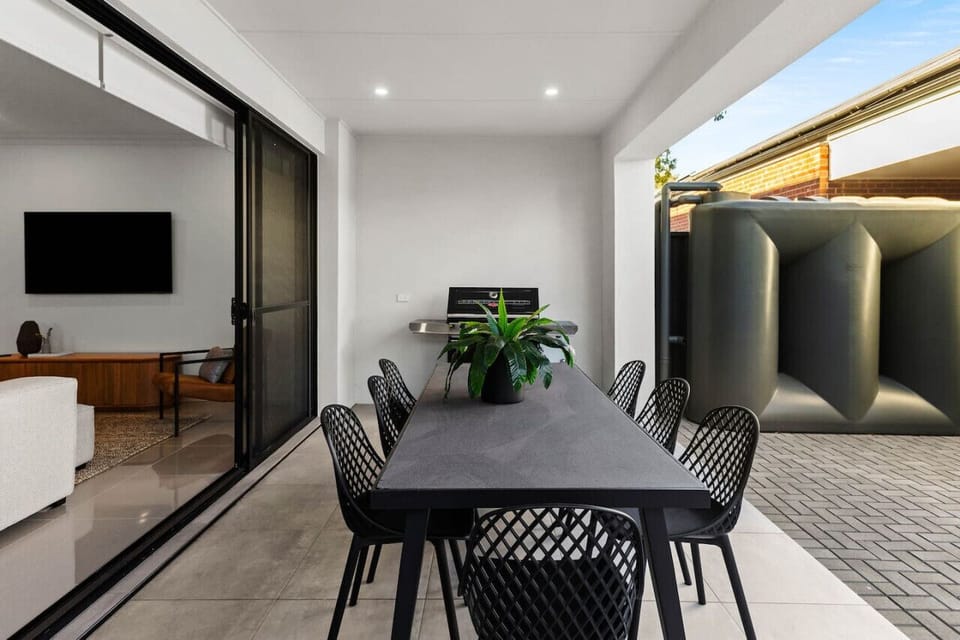 Modern outdoor dining area with a black table and matching chairs, a potted plant on the table, and a view through sliding doors to an indoor space with a TV and furniture. There is also a large water tank visible in the outdoor area.