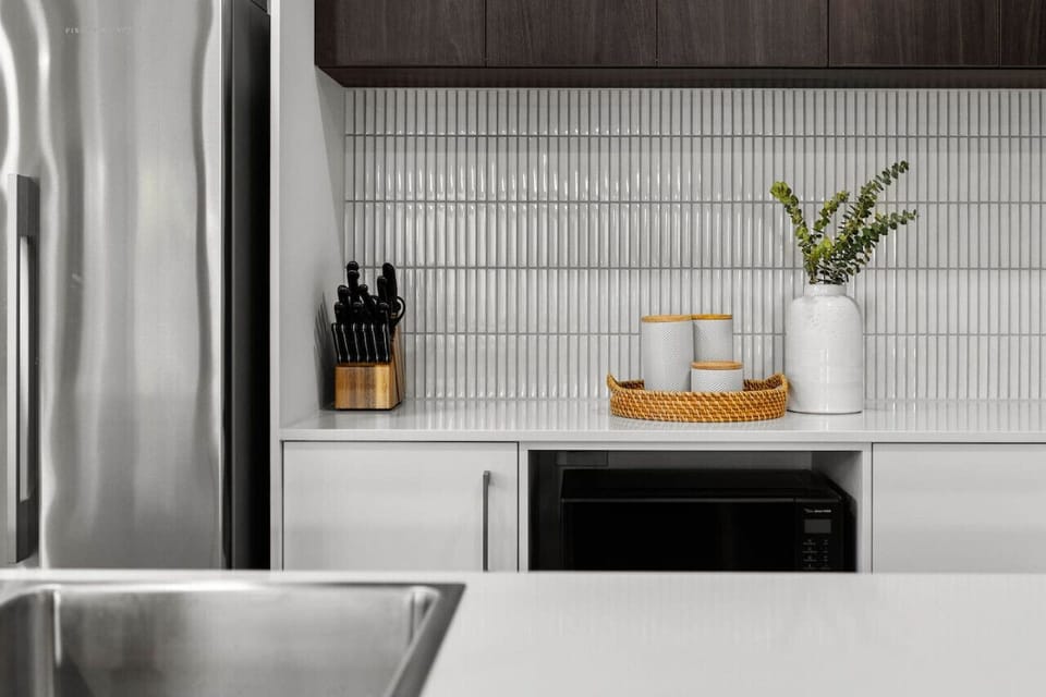 A modern kitchen counter with a sleek design, featuring a knife block, decorative canisters on a wicker tray, a white vase with greenery, and a stainless steel refrigerator in the background, all set against a backdrop of stylish vertical tiled wall.