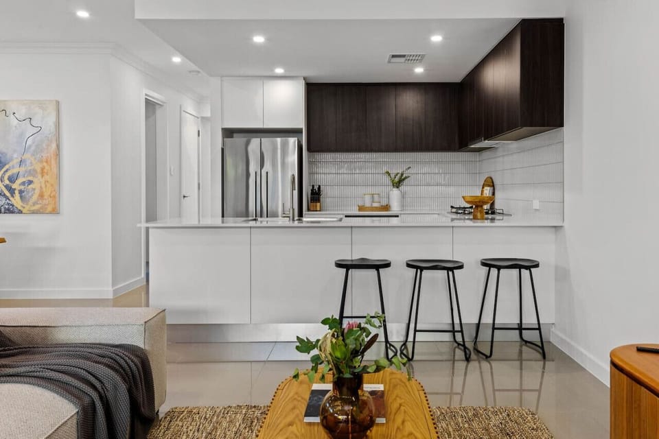 A modern, open-plan living space with a sleek kitchen area featuring white countertops and dark wooden cabinets. There are three black stools at the kitchen counter, and the adjacent living area includes a sofa with a throw blanket and a wooden coffee tab