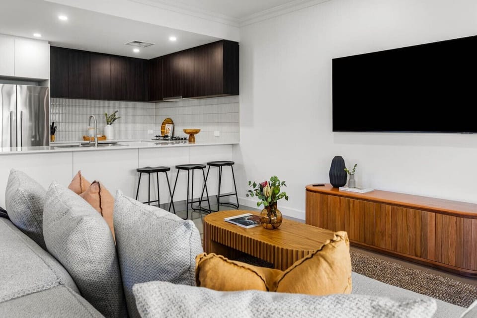 The foreground showcases a cozy living room with a large TV mounted on the wall, a wooden coffee table adorned with a vase of plants, and comfortable gray and orange cushions on the sofa.