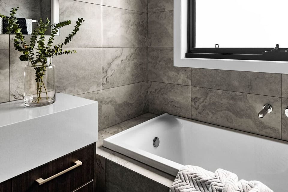 A modern bathroom with a sleek design. It features a white bathtub, a white countertop with a dark wooden cabinet, and grey marble tiles on the walls. A vase with green foliage adds a touch of nature, and a window allows natural light to illuminate the sp