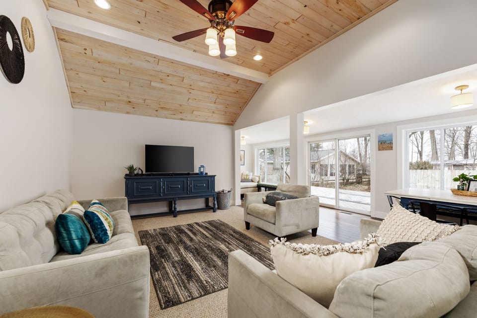 Our living room is bright and spacious, with modern furniture, a wooden ceiling, and large windows showcasing the outdoor view.