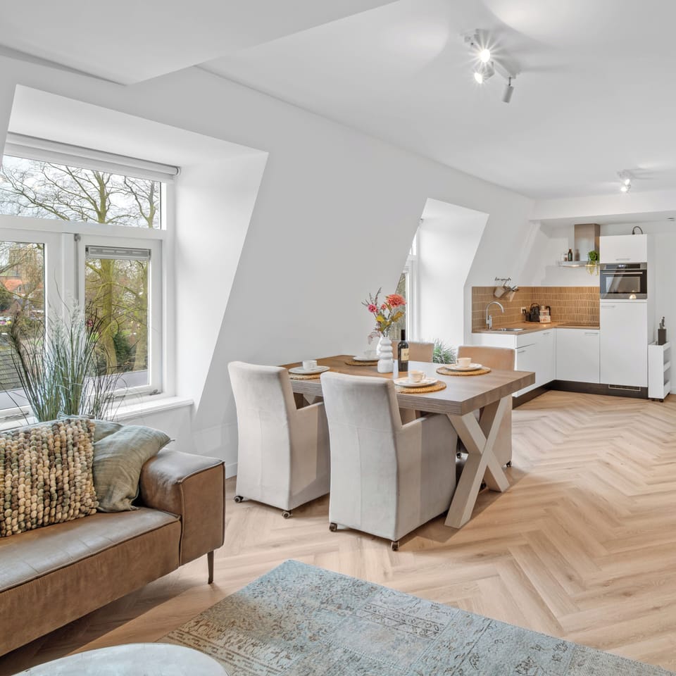 A stylish open-concept interior with a beige sectional, rustic wooden dining table, and large windows showcasing an inviting, airy atmosphere.