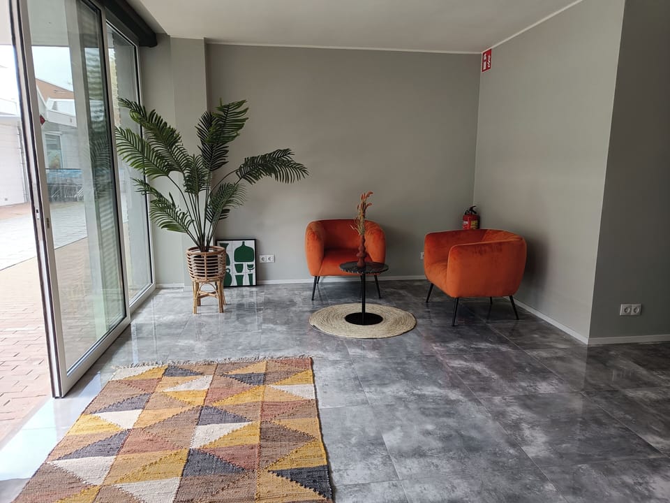 A spacious seating area near the entrance is brightened by large windows, featuring a blend of vibrant orange chairs, greenery, and a patterned rug, offering a perfect spot for relaxation or welcoming guests.