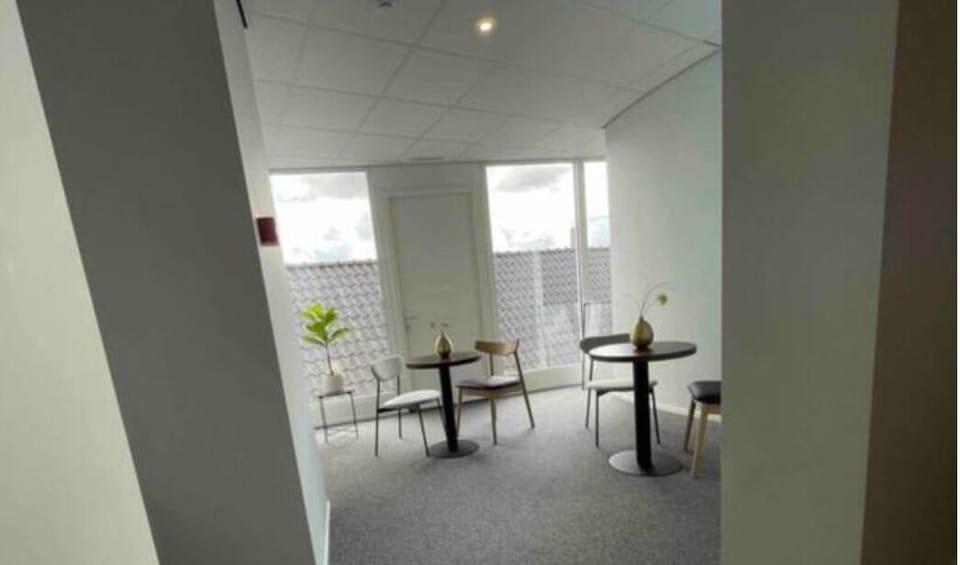 
Bright lounge area with round black tables, simple wooden and metal chairs, a potted plant, and large windows offering natural light and rooftop views.