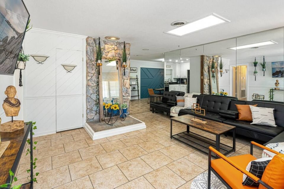 A cozy living room with a modern design featuring a large, dark sofa with accent pillows, a mirrored wall creating an illusion of spaciousness, and stone decor elements around a fireplace. The room has light-colored tiled flooring and is accented with vibrant touches like a bright orange chair and decorative plants. The space extends to an open kitchen area visible in the background.