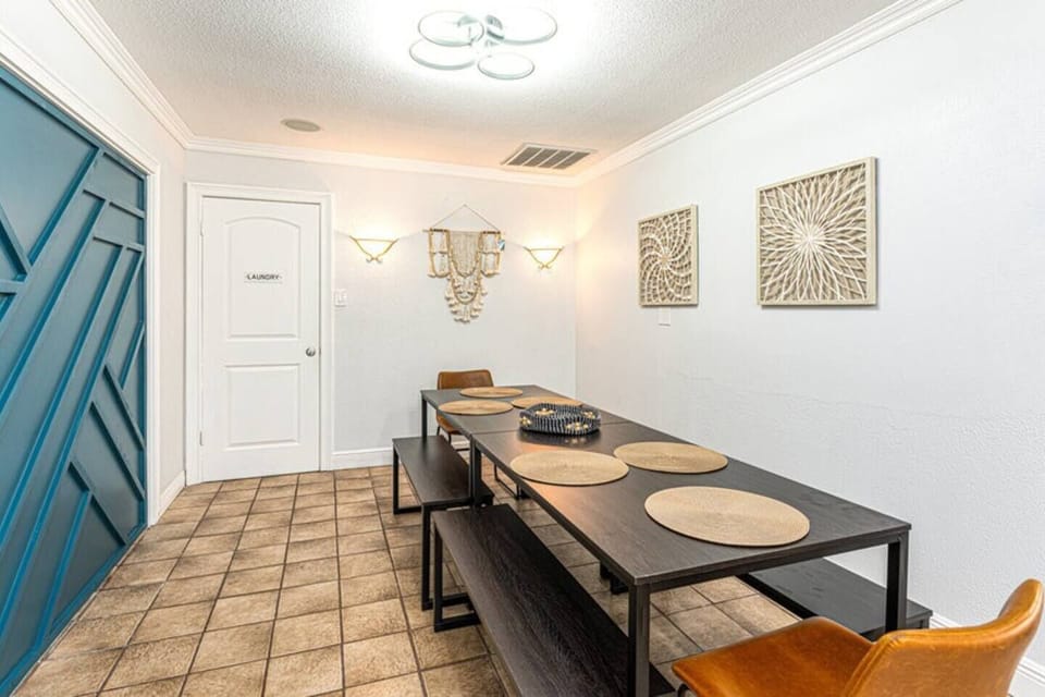 A stylish dining room featuring a long wooden table with matching benches and chairs. The table is set with round woven placemats, giving a warm and inviting atmosphere. On the right wall, there are decorative wall hangings with intricate designs, while modern sconces provide soft lighting. The room has tiled flooring and a blue accent wall on the left that adds a pop of color....